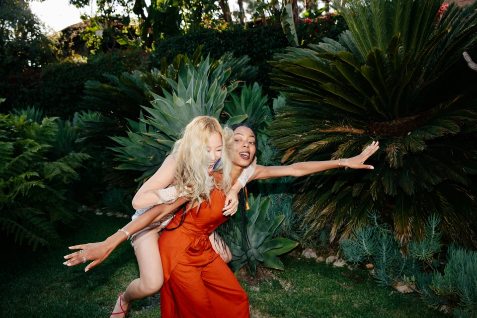 Two women pose outdoors amid lush greenery: one carries the other piggyback style with arms outstretched, in an expressive moment.
