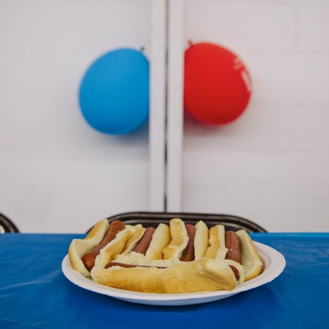 Hot dogs on a plate sitting on a blue tablecloth, with red and blue balloons in the background, food photography for catering menu.