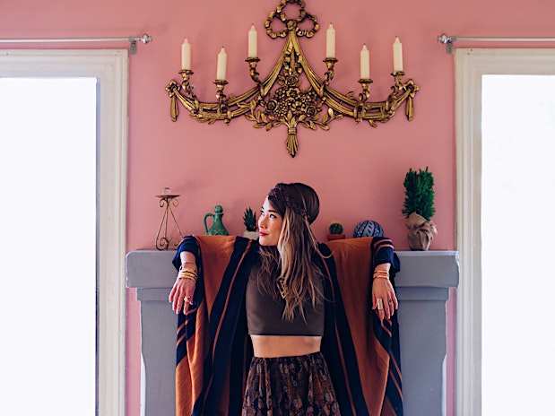 A woman with a head wrap and brown crop top stands in front of a gray fireplace with a vintage gold chandelier on a pink wall.