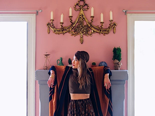 A woman with a head wrap and brown crop top stands in front of a gray fireplace with a vintage gold chandelier on a pink wall.
