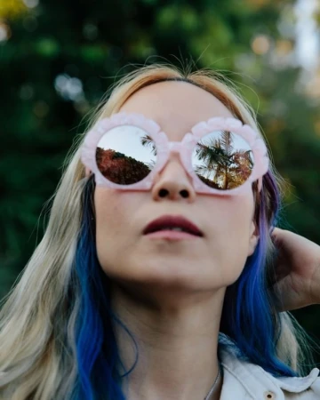Woman wearing oversized pink round sunglasses with reflective lenses mirroring palm trees against an out-of-focus green background