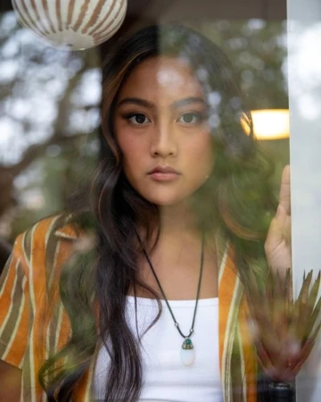 Close-up portrait of a young woman with dark wavy hair wearing a striped shirt and pendant necklace photographed through a glass window.