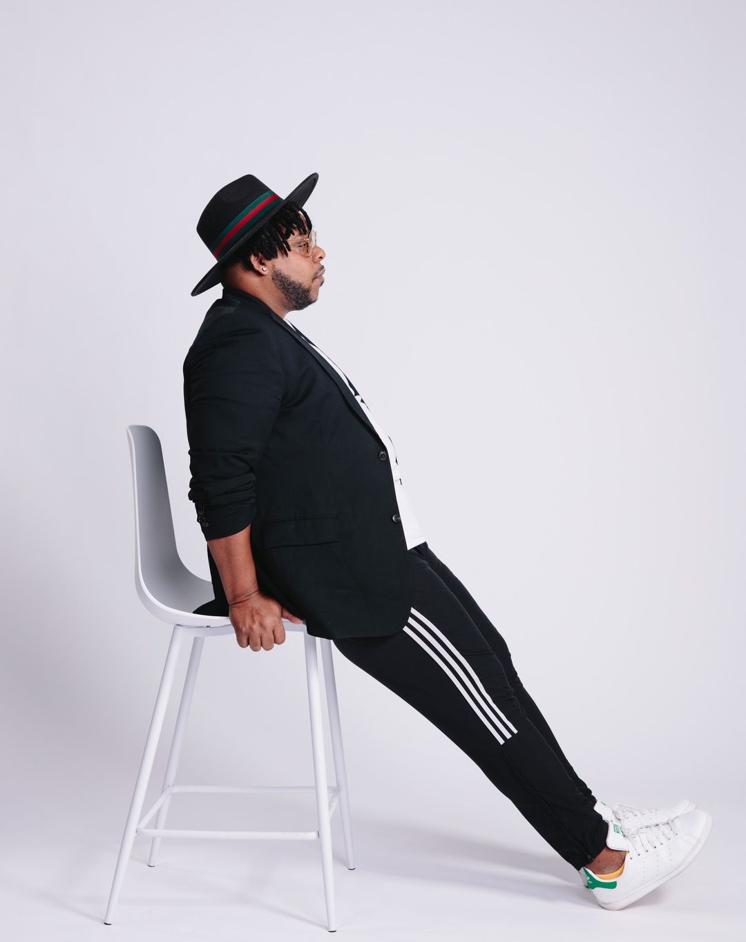Man wearing a black fedora, blazer, and striped track pants leaning on a white stool against a white seamless background