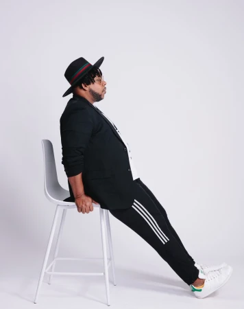 Man wearing a black fedora, blazer, and striped track pants leaning on a white stool against a white seamless background