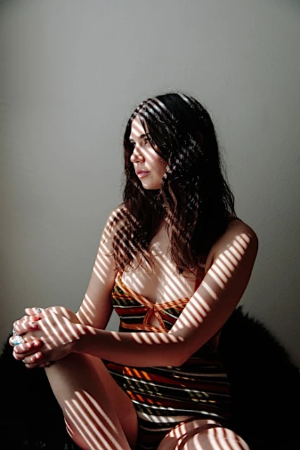 Portrait of a young woman in striped swimsuit sitting in room with window light creating striped shadows on skin and white wall.