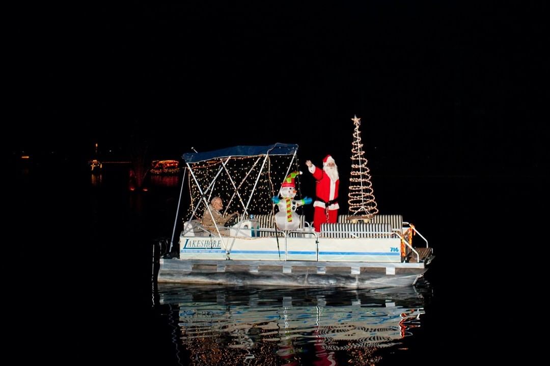 Holiday boat parade on a calm river at night featuring Santa Claus, a snowman, and a decorated Christmas tree on a pontoon boat.