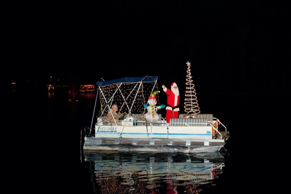 Holiday boat parade on a calm river at night featuring Santa Claus, a snowman, and a decorated Christmas tree on a pontoon boat.
