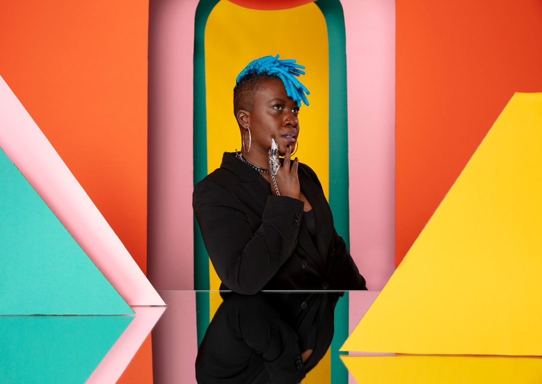 Studio portrait of an adult woman with blue dreadlocks, wearing a black blazer, posing with multicolored geometric shapes in the background.