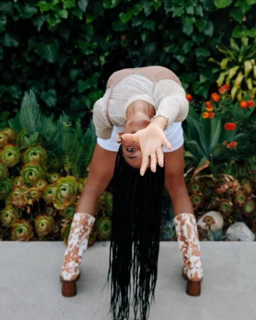 Woman in white shorts and cow print boots bending backwards in garden with lush green foliage, red flowers, and succulent plants.