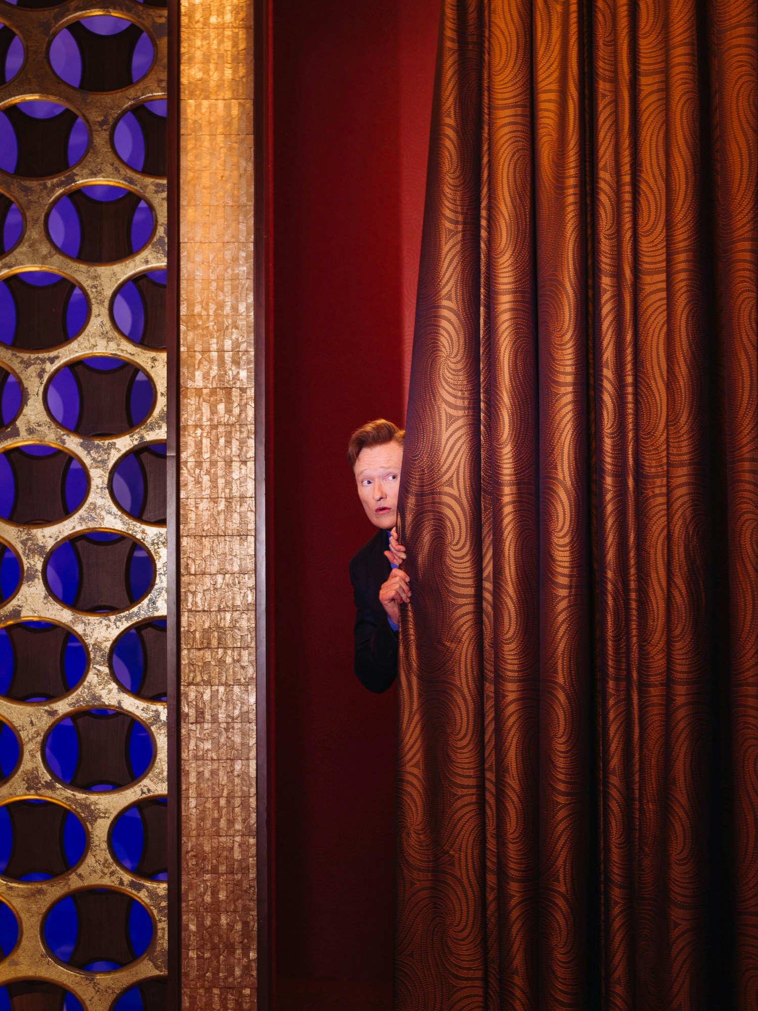 Man peeking from behind a heavy patterned curtain next to a decorative panel with circular cutouts