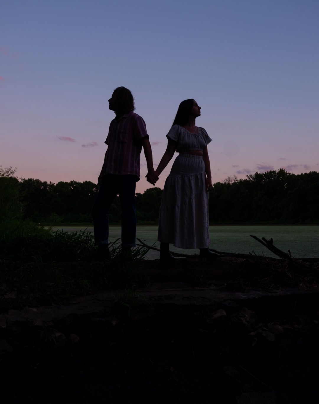 Silhouette of a man and a woman holding hands at dusk, standing in front of a marsh and trees in a dark, natural landscape.
