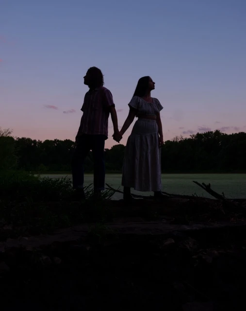Silhouette of a man and a woman holding hands at dusk, standing in front of a marsh and trees in a dark, natural landscape.