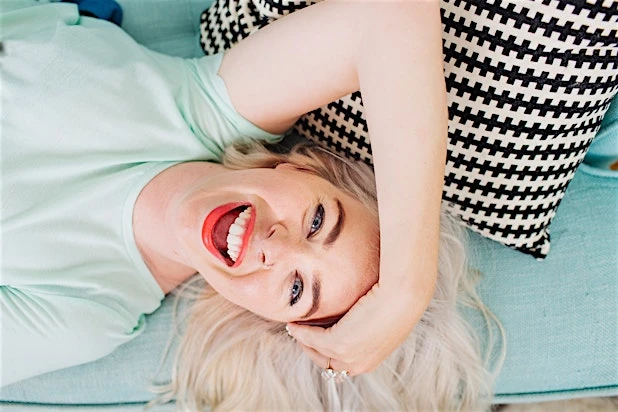 Smiling fair-skinned woman with red lipstick and blonde hair poses in a bright studio with a black and white pillow behind her.