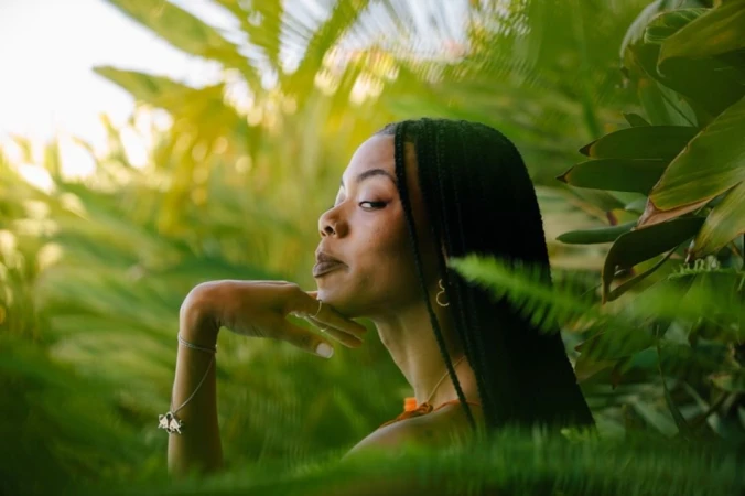Portrait of a Black woman with braided hair resting her chin on her hand amidst lush green tropical foliage by Brinson Banks.