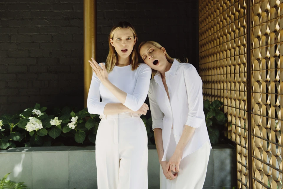 Two adult females pose in front of a dark brick wall and gold decorative panel, wearing all-white outfits in an indoor setting.