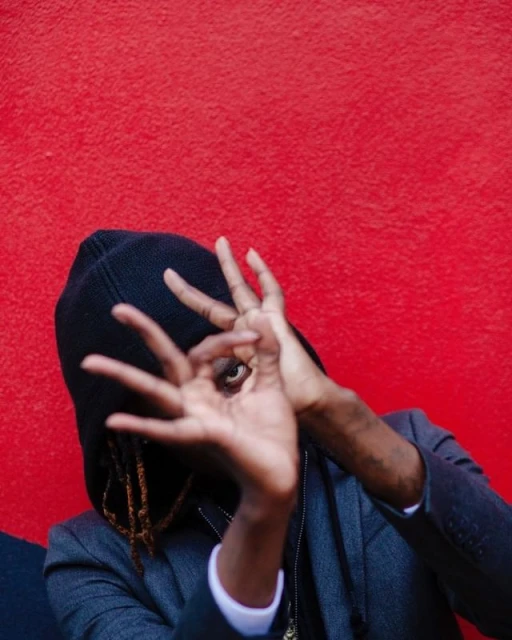 Person wearing a hooded jacket makes a circle with their hands, showing their eye, against a solid red background. Medium shot.