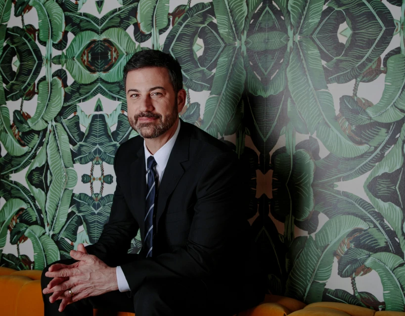 Portrait of Jimmy Kimmel in a black suit, seated against a background of tropical leaves, with a direct gaze and hands clasped.
