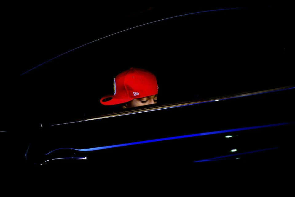 Person in a red baseball cap is visible from the shoulders up inside of a dark vehicle, with a dark background.