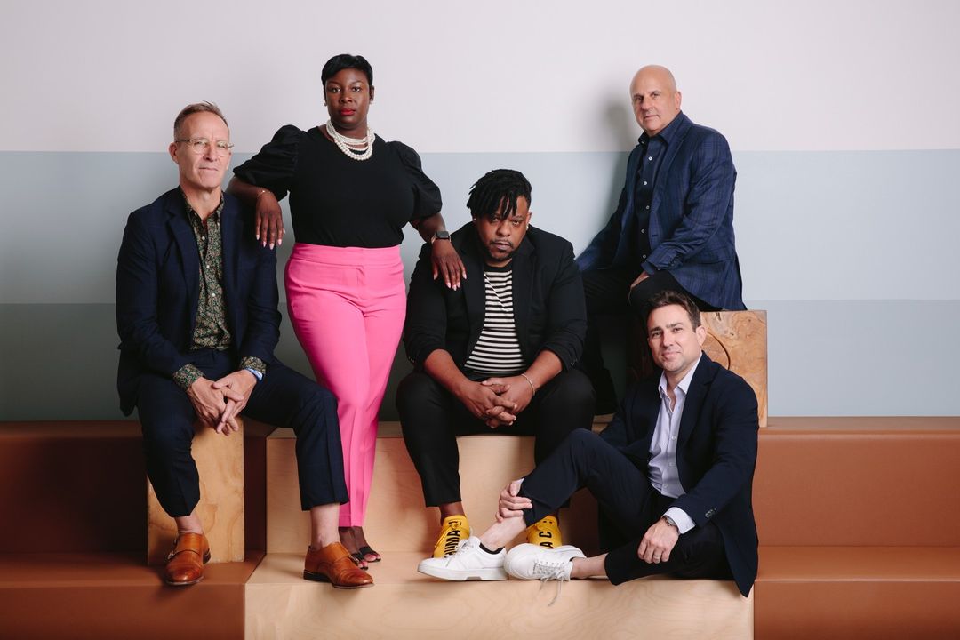 Group portrait of five adults seated indoors on wooden blocks. The diverse group wears fashionable business casual attire with jackets.