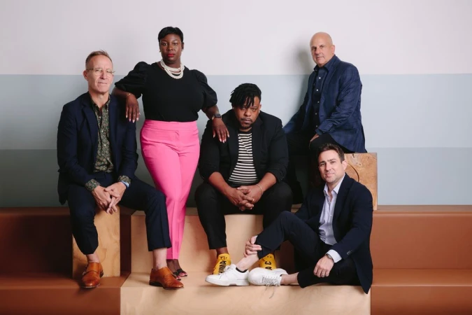 Group portrait of five adults seated indoors on wooden blocks. The diverse group wears fashionable business casual attire with jackets.