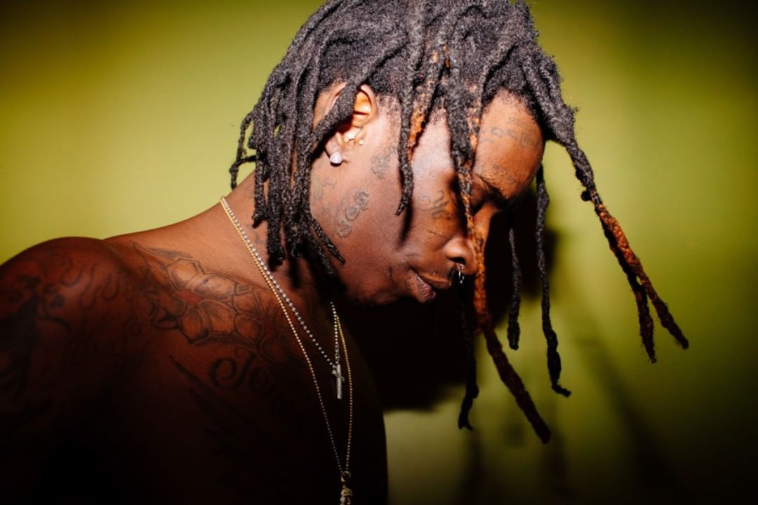Adult man with dreadlocks and tattoos gazes downward against a chartreuse backdrop in a close-up studio shot with natural-looking lighting.