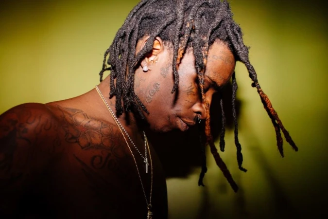 Adult man with dreadlocks and tattoos gazes downward against a chartreuse backdrop in a close-up studio shot with natural-looking lighting.