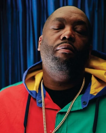 Portrait of an adult Black man with eyes closed, wearing a colorful jacket and gold chains, against a blurred blue backdrop.