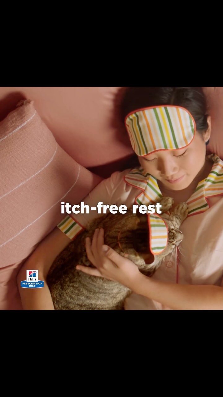 Adult woman and tabby cat snuggle in a cozy pink bed. Both wear colorful striped sleep masks in this adorable, intimate scene.
