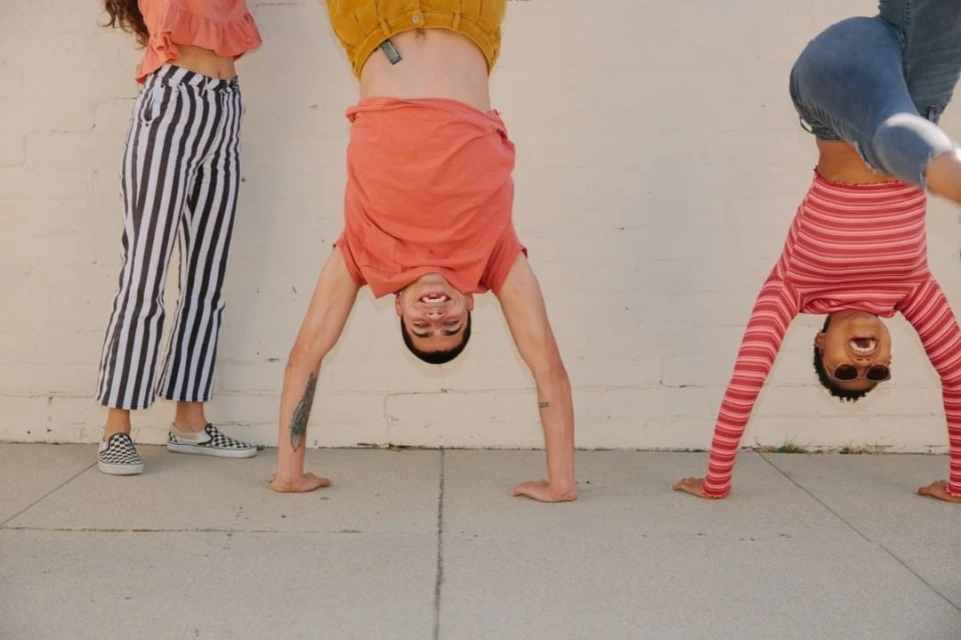 Three young adults in handstand poses against a white brick wall on a concrete surface wearing casual clothing in the daytime.