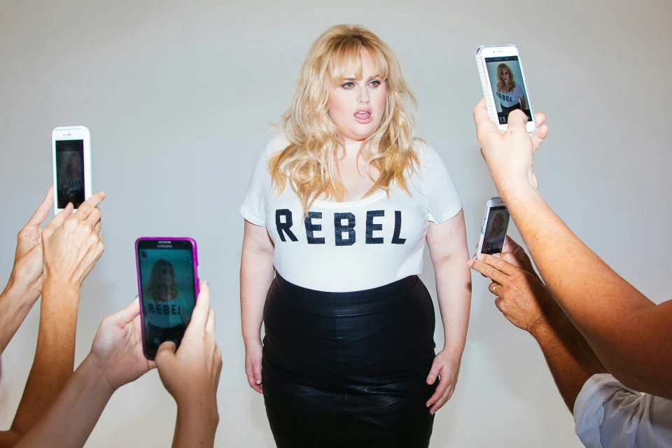 Blonde woman in a black leather skirt and white t-shirt with 'REBEL' on it is surrounded by smartphones taking photos of her.