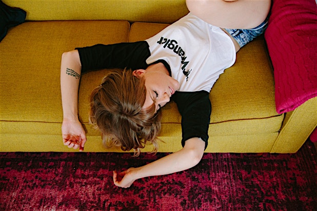 Woman lying upside down on a yellow sofa with a red pillow, wearing a Wrangler t-shirt, in an indoor setting with a patterned red rug.