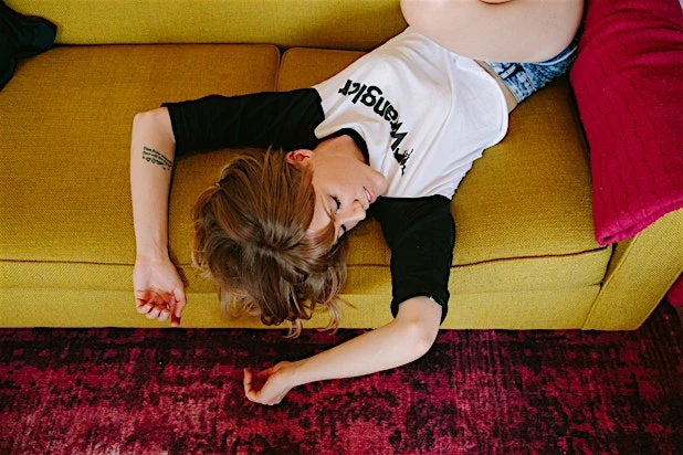Woman lying upside down on a yellow sofa with a red pillow, wearing a Wrangler t-shirt, in an indoor setting with a patterned red rug.