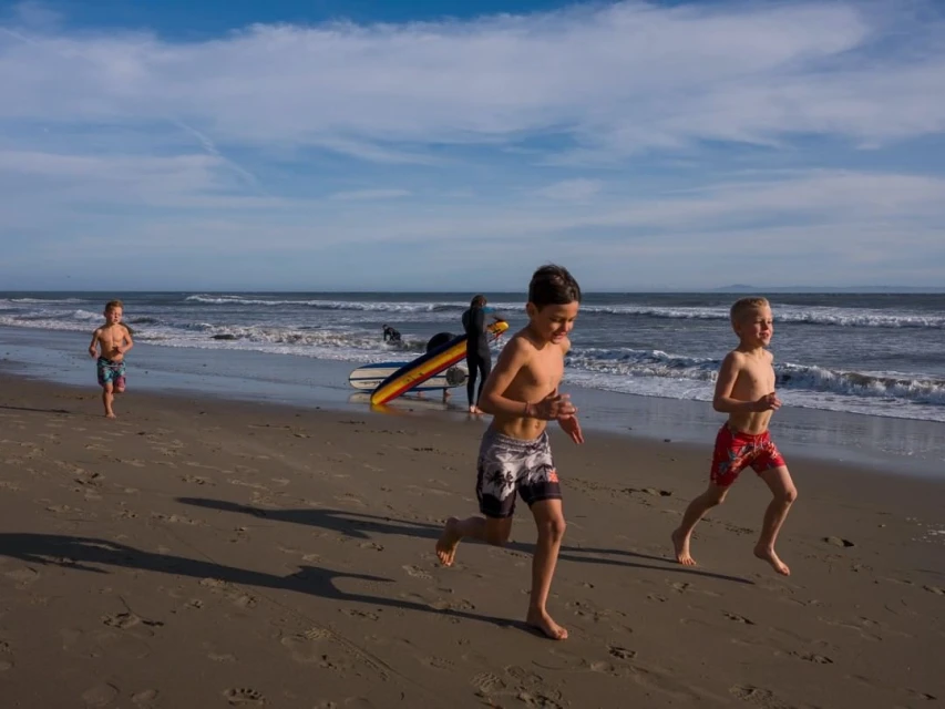 Three young boys are running barefoot on a sandy beach in the sunlight. A surfboard and waves are visible in the background.
