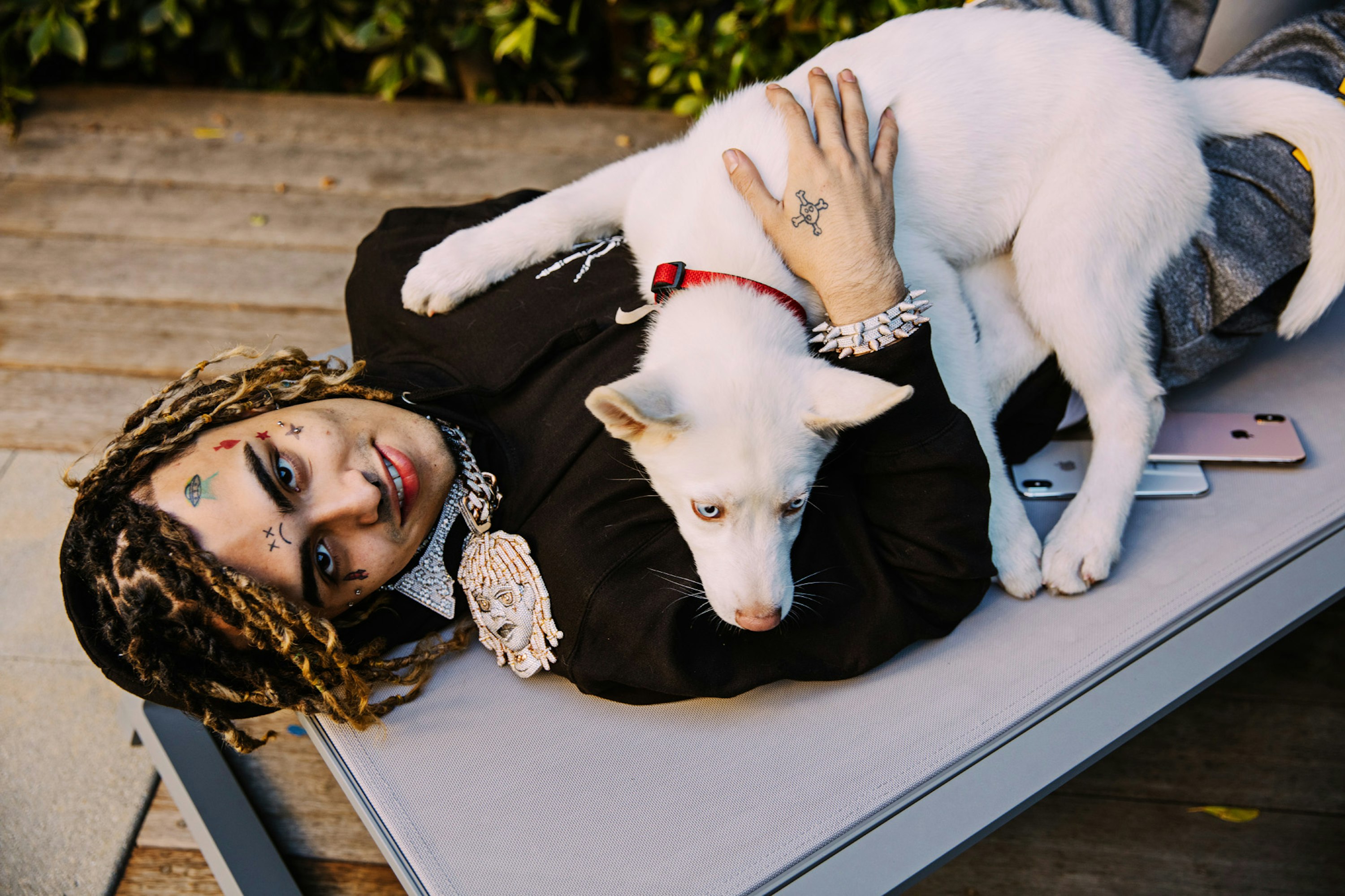 Rapper lays on lounger with a white dog. Man has tattoos and dreadlocks, wearing jeweled necklace and black jacket outdoors.