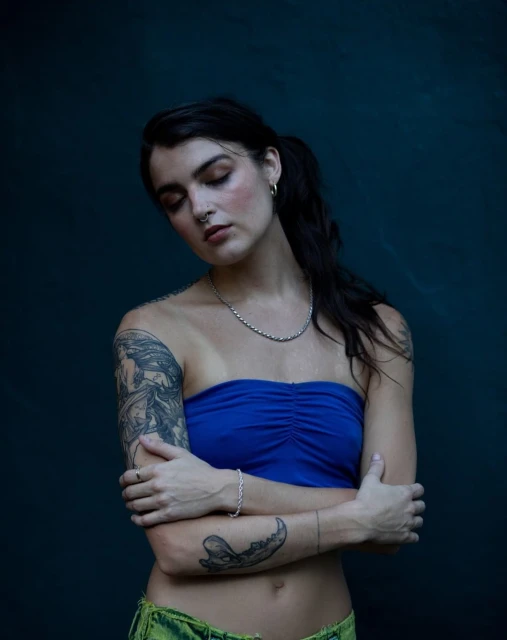 Close-up studio portrait of a woman with dark hair and tattoos, wearing a blue tube top and green bottoms against a dark backdrop.