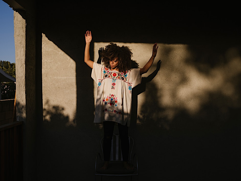 Woman in embroidered tunic stands on chair outdoors against wall with sunlight and shadows, arms raised, wearing dark pants.