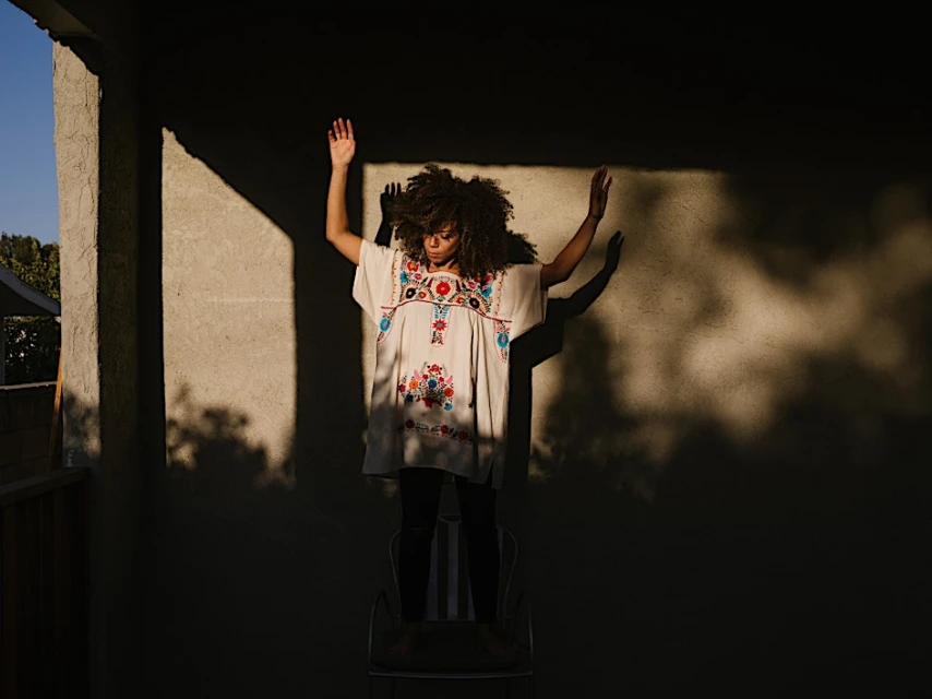 Woman in embroidered tunic stands on chair outdoors against wall with sunlight and shadows, arms raised, wearing dark pants.