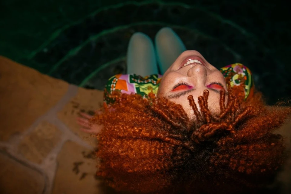 Woman with red curly hair and vibrant makeup relaxes in a pool, smiling and looking up towards the camera in a close-up shot.