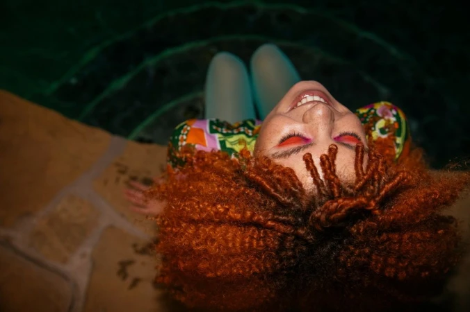 Woman with red curly hair and vibrant makeup relaxes in a pool, smiling and looking up towards the camera in a close-up shot.