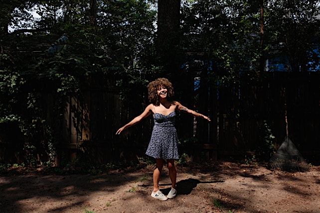 A young woman with curly hair stands in a sunny backyard wearing a floral dress, smiling with arms outstretched, surrounded by greenery.