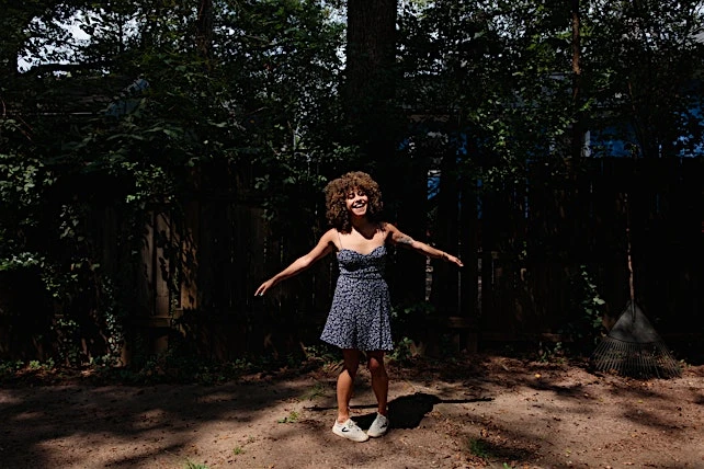 A young woman with curly hair stands in a sunny backyard wearing a floral dress, smiling with arms outstretched, surrounded by greenery.