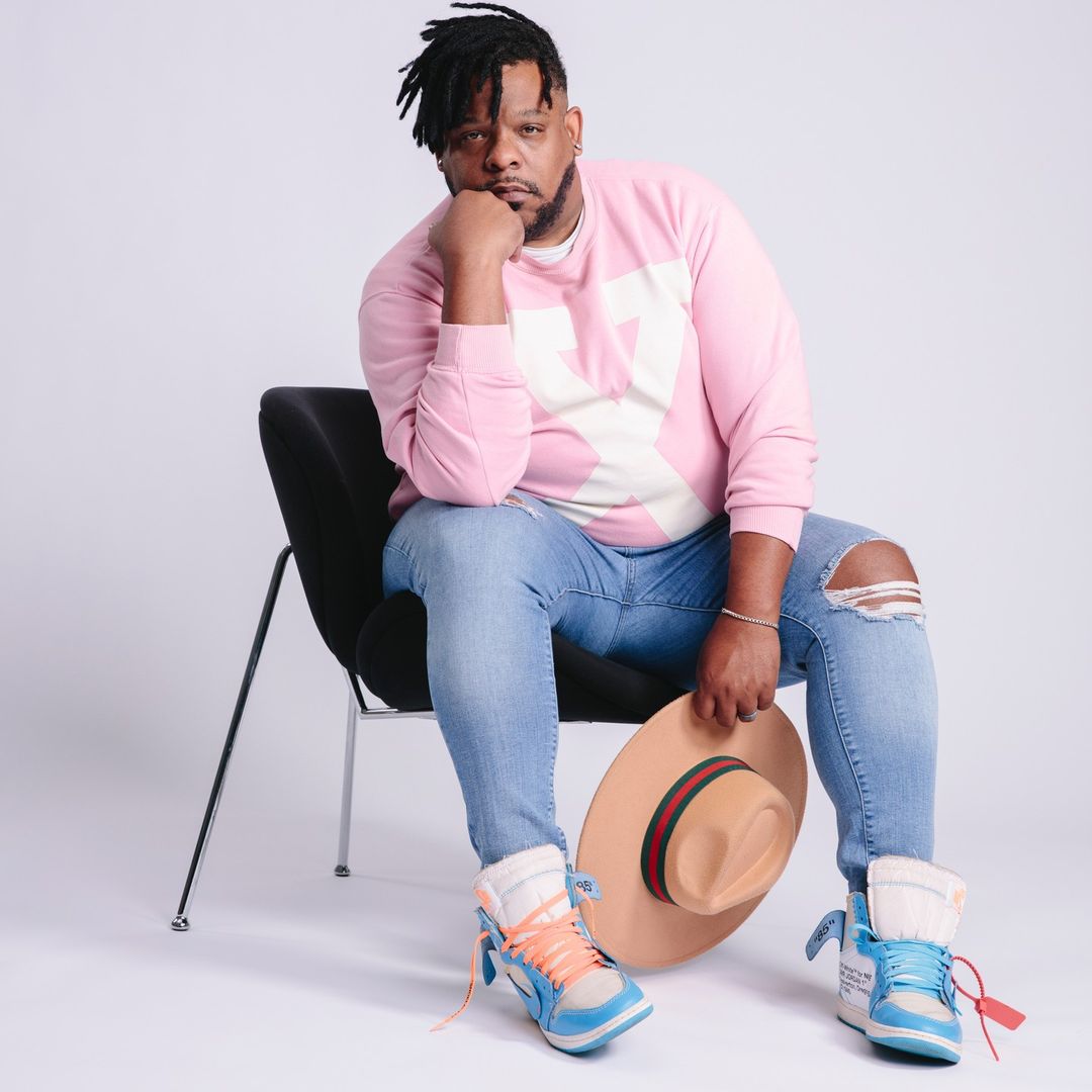 Man in pink sweatshirt sits in chair holding a tan hat, wearing light blue jeans and matching athletic shoes against white background.