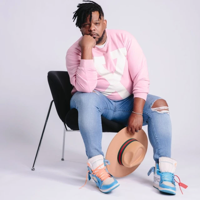 Man in pink sweatshirt sits in chair holding a tan hat, wearing light blue jeans and matching athletic shoes against white background.