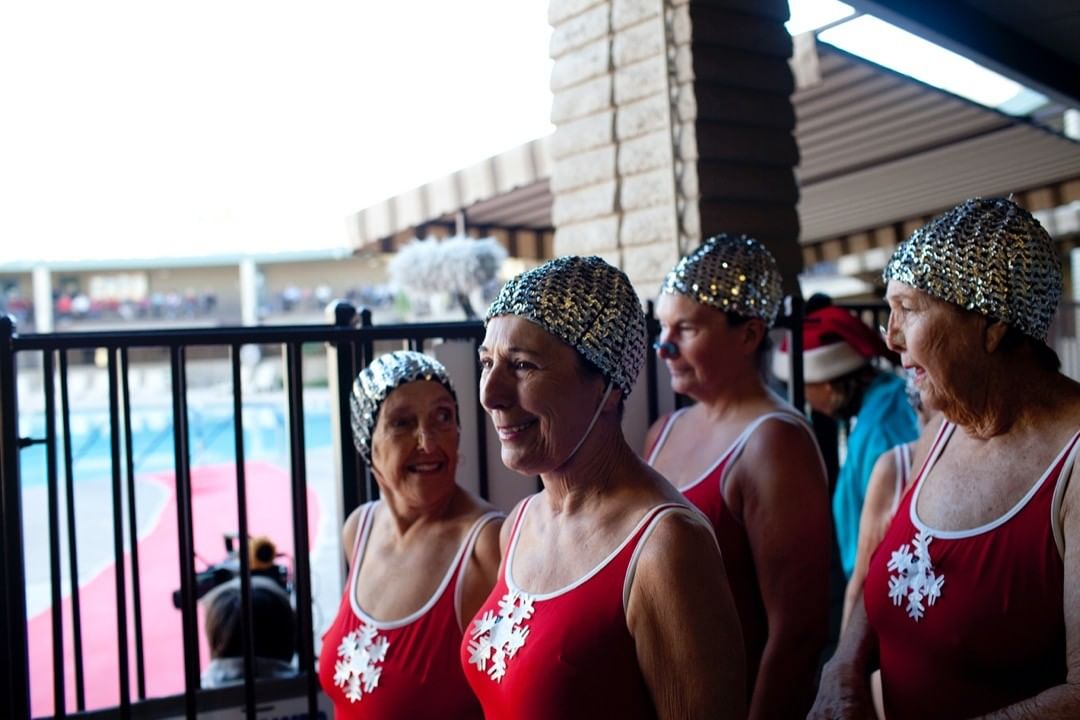 Group of smiling senior women in matching red swimsuits and silver swim caps, standing by a pool with a building in the background.