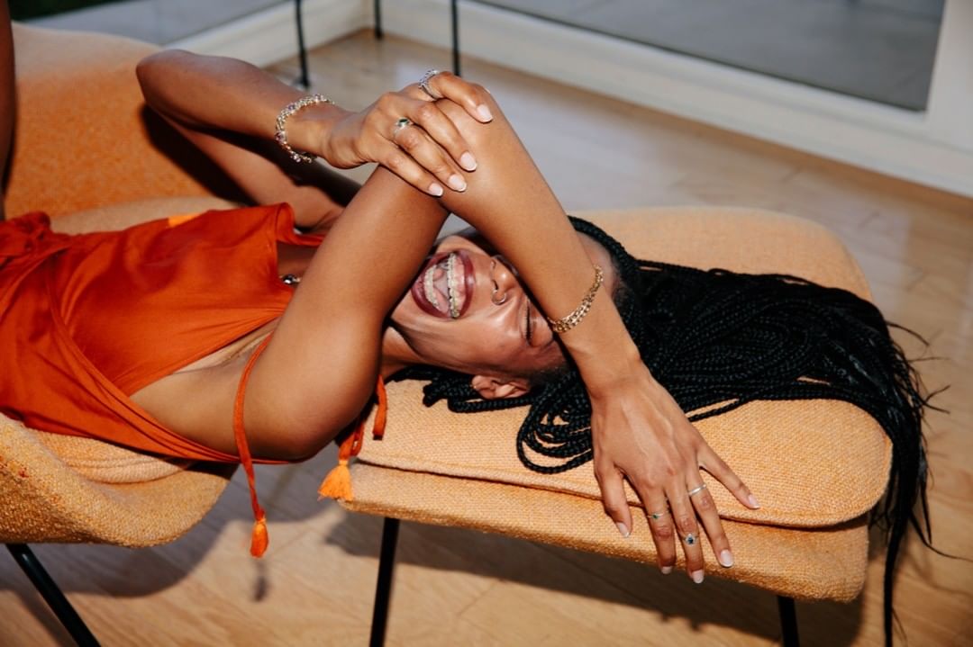 A happy black woman with braids lies on an orange chair with arms crossed, wearing an orange top and gold bracelets, smiling widely.
