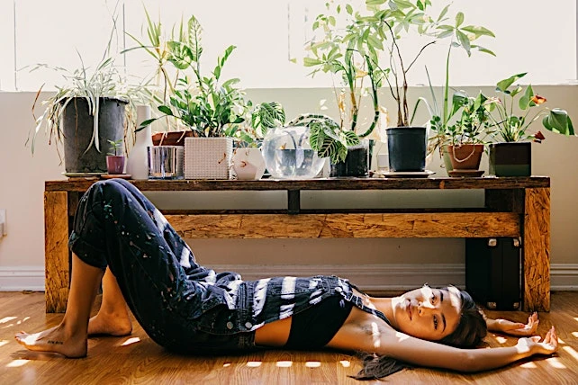 Young woman in overalls lies on wooden floor with houseplants on shelf. Bright sunlight indoors, casual fashion, wellness lifestyle concept.