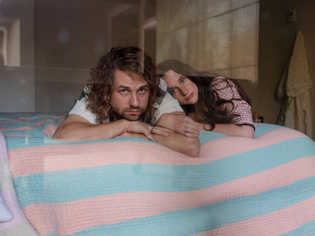 A fair-skinned man and woman are lying on a striped pastel bedspread, with the man in the foreground and the woman behind him.