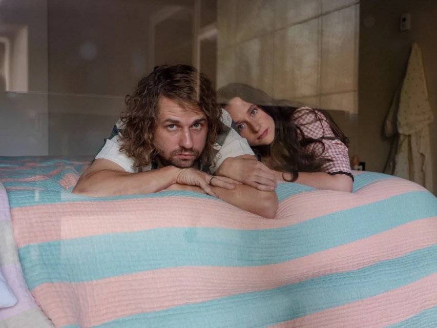 A fair-skinned man and woman are lying on a striped pastel bedspread, with the man in the foreground and the woman behind him.