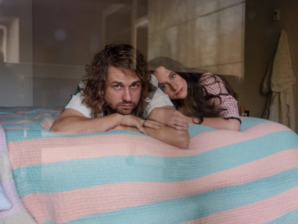 A fair-skinned man and woman are lying on a striped pastel bedspread, with the man in the foreground and the woman behind him.