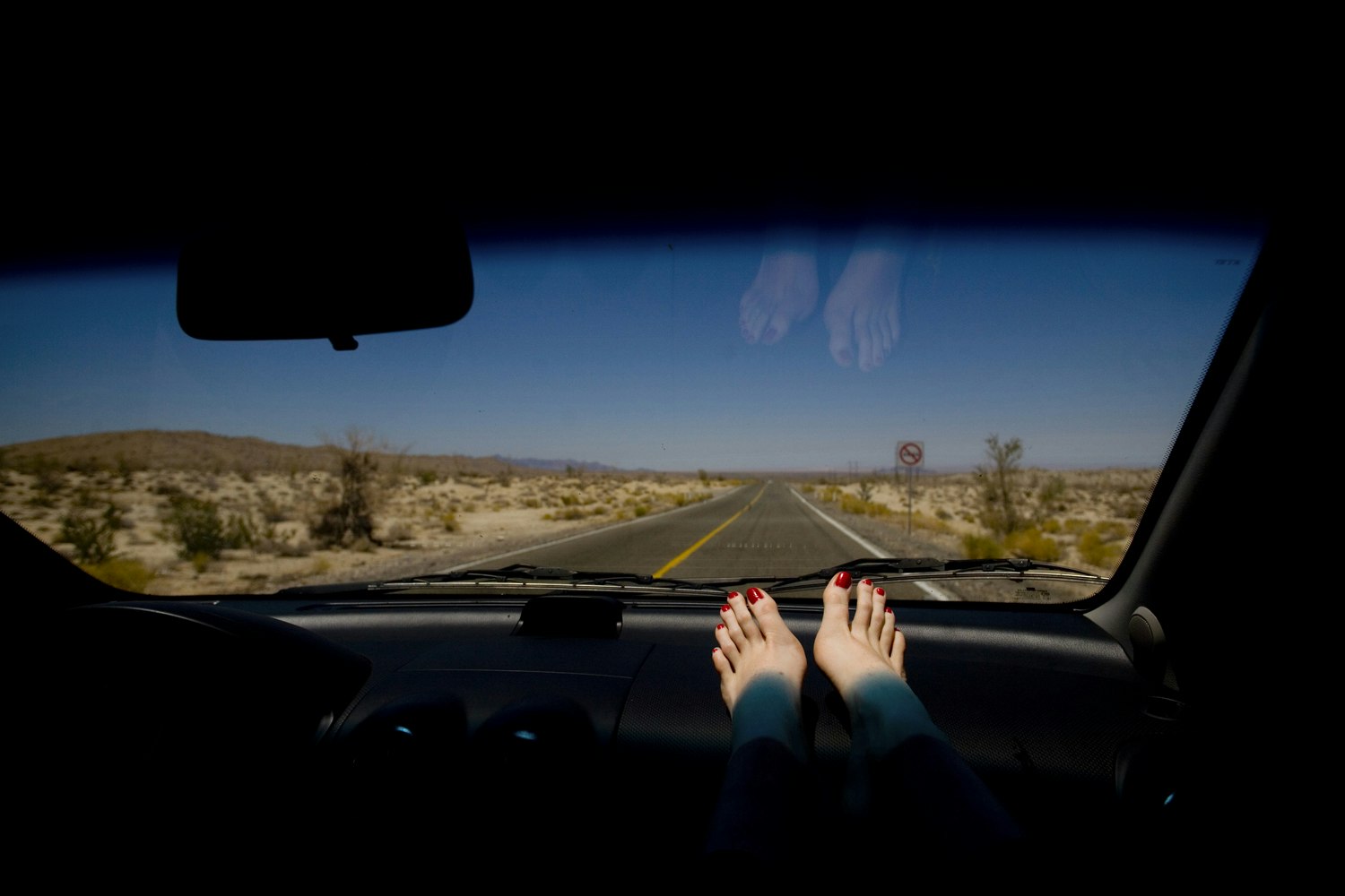 Feet with red nail polish resting on a car dashboard facing an empty desert highway